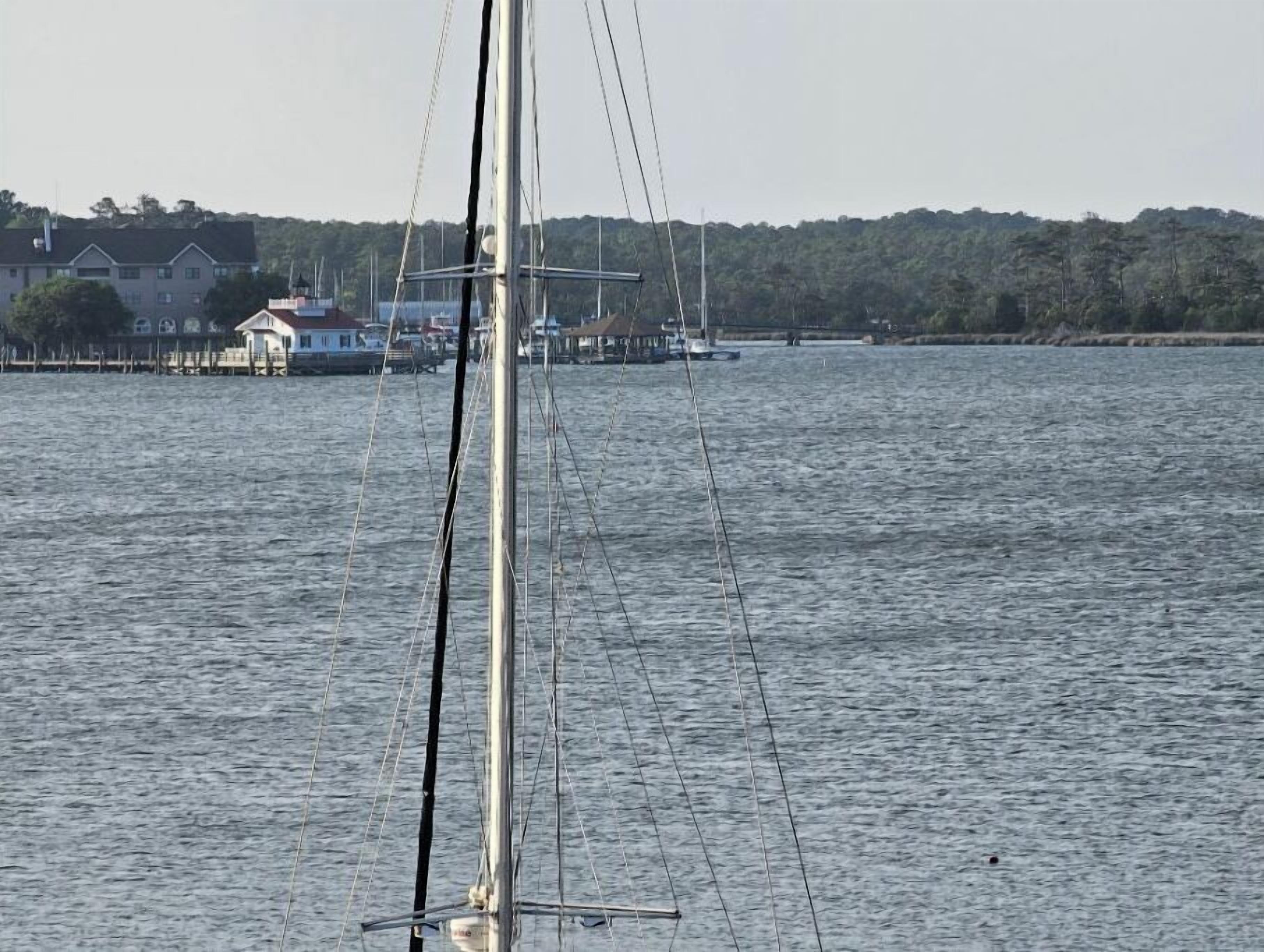 Un velero con mástil y aparejo, un muelle con un edificio, y una orilla boscosa al fondo.