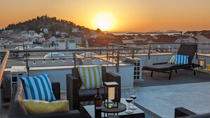 A rooftop terrace with outdoor seating, a table with drinks, and a view of a sunset over a cityscape.