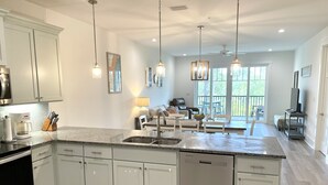A modern kitchen with white cabinets, a dishwasher, and a coffee maker. There is a dining area with a table and chairs, and a living room with a sofa and a TV.