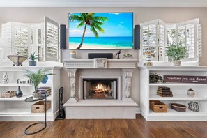 A living room with a fireplace, a television displaying a beach scene, and open shutters.