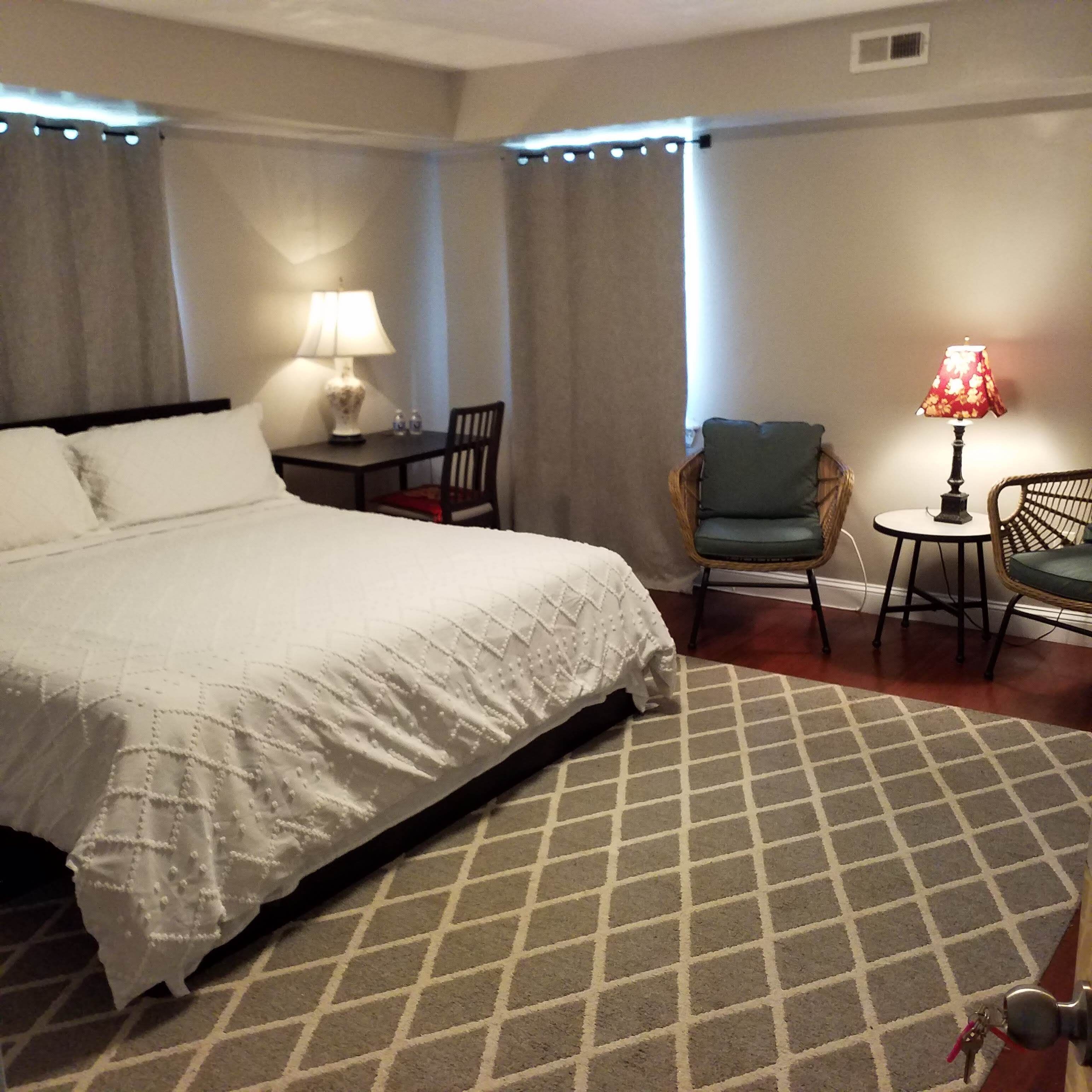 Habitación de hotel con una cama grande, una mesita con una lámpara, una silla y otra silla con cojín.