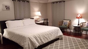 A neatly made bed with a white duvet, a bedside table with a lamp, and a chair with a cushion. - Fairmount House at The Mann (Philadelphia)