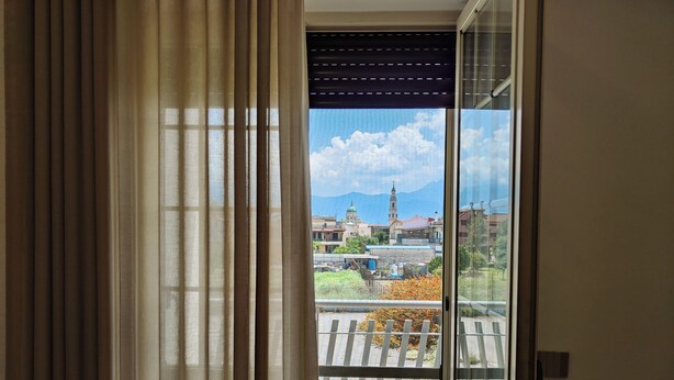 A view through a window with curtains, revealing a balcony with a tiled floor and a railing.