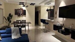 Uma sala de estar moderna com um sofá azul, mesa de jantar e cadeiras. Há uma TV na parede e um pequeno bar com TV e objetos decorativos.