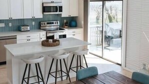 Una cocina moderna con armarios blancos, una isla central con taburetes, una mesa de comedor de madera y un amplio ventanal corredera que da a una terraza.