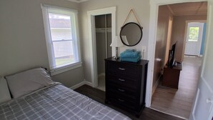 Un dormitorio con cama, cómoda y espejo. Hay una ventana con persianas y una puerta que da a otra habitación.