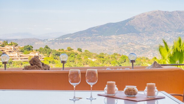 Ein Tisch mit zwei Weingläsern und Kerzen, mit Blick auf eine gebirgige Landschaft und Gebäude in der Ferne.