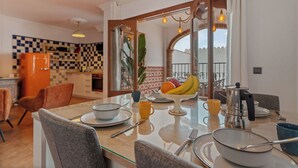 A dining area with a glass table, chairs, and a fruit bowl. A kitchen with checkered tiles and a refrigerator is visible in the background. - casa carpa andalucia II (Nerja)