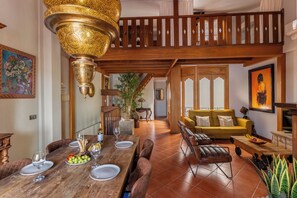 Comedor con una mesa de madera dispuesta para seis comensales, una lámpara colgante ornamentada y una escalera que sube a un nivel superior.