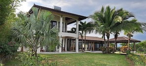 Une maison à deux étages dotée d’un patio couvert, entourée de plantes tropicales et de palmiers.