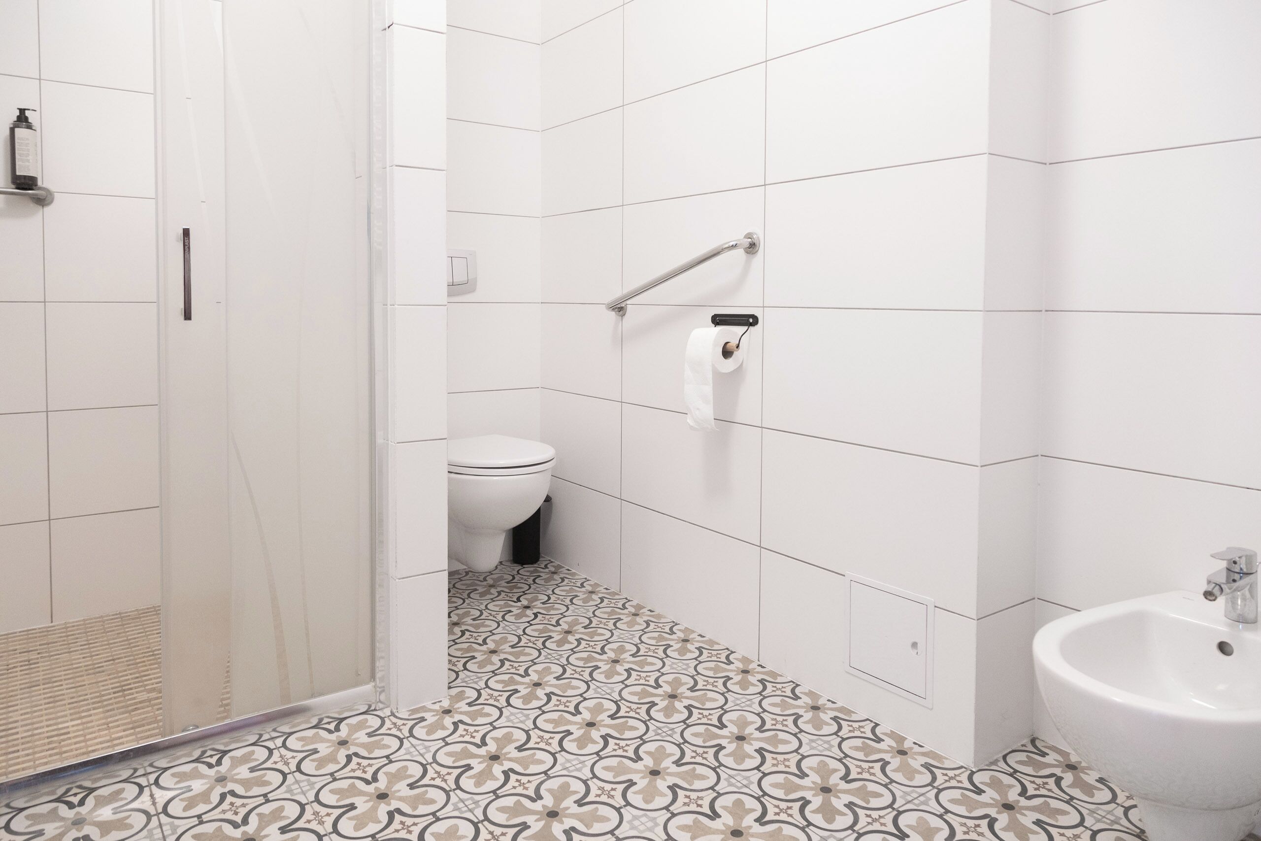 Un baño con azulejos blancos, piso estampado, inodoro, bidé y zona de ducha.
