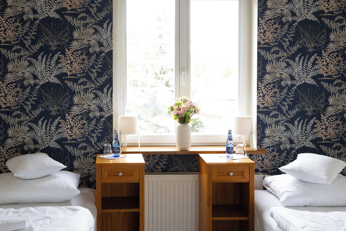 A hotel room with a large window, a desk with a vase of flowers, and two beds with white linens.
