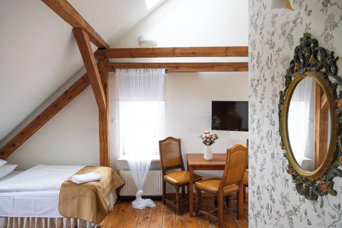 A room with a sloped ceiling, wooden beams, a bed, a table with chairs, a mirror, and a window with sheer curtains.