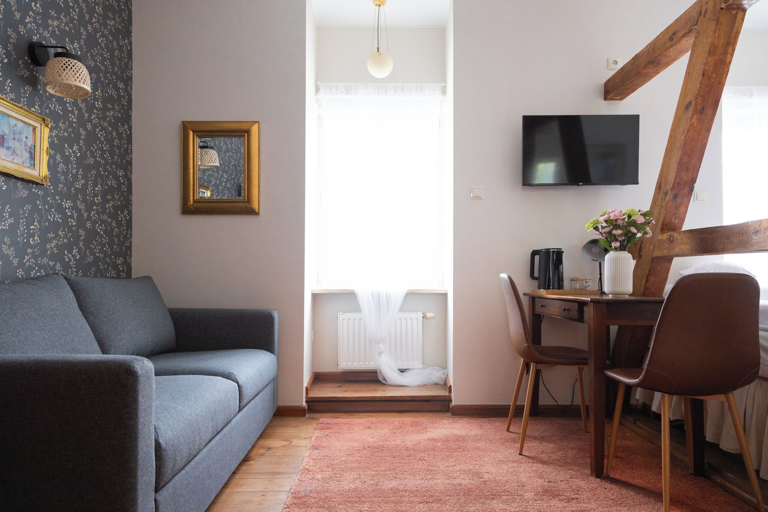 Un salon moderne avec un canapé gris, une table à manger en bois accompagnée de deux chaises, un téléviseur fixé au mur et une fenêtre ornée de rideaux légers.
