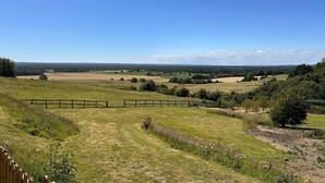 Ett lantligt landskap med en inhägnad åker, ett trästaket och en avlägsen vy över böljande kullar under en klar himmel.