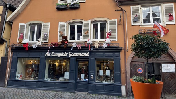 A building with a storefront labeled 'Le Comptoir Gourmand' featuring a large window display, a balcony with various decorations, and a potted plant. - Zum Storike Nescht (Barr)