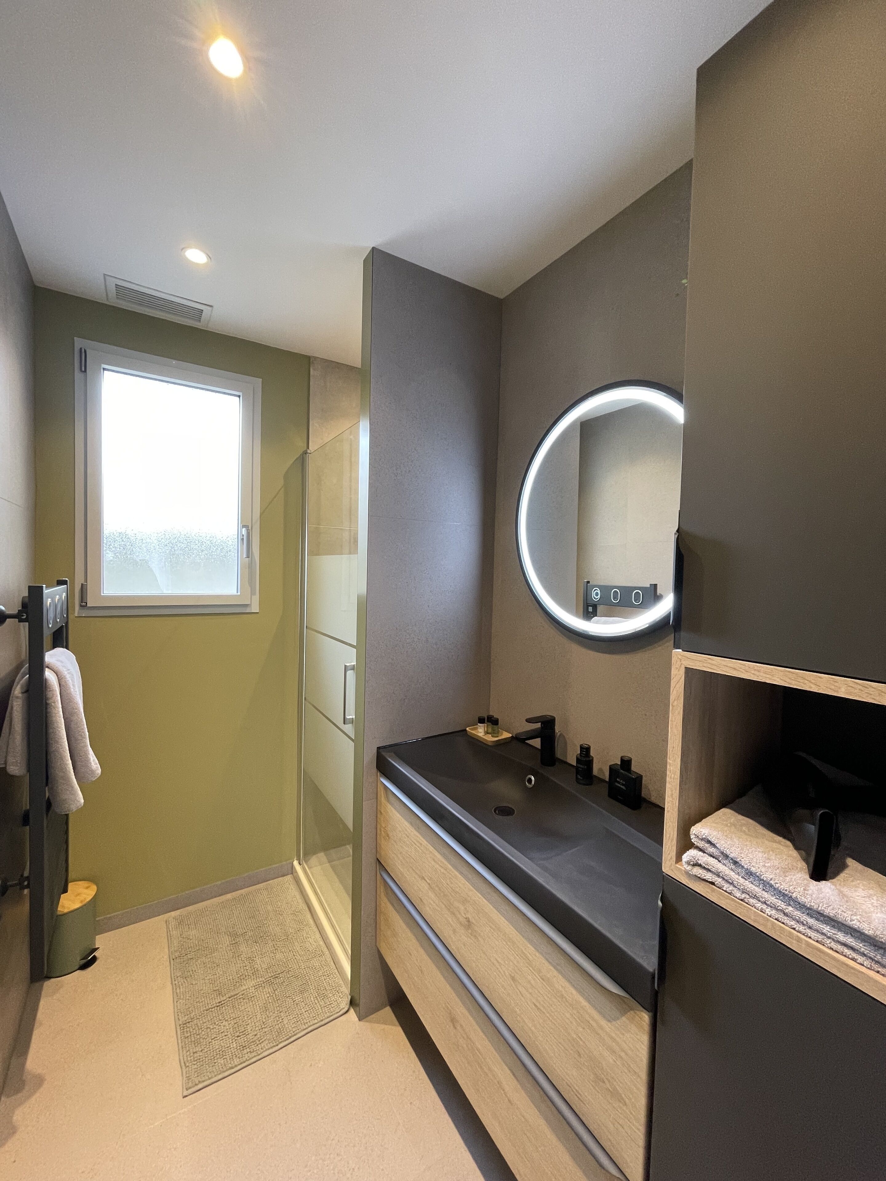 Ein modernes Badezimmer mit Waschtisch, rundem Spiegel mit Beleuchtung, Handtuchhalter und Fenster.