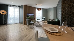 Una sala moderna con suelo de madera, un televisor montado en la pared, una zona de comedor con una mesa para dos personas y un sofá.