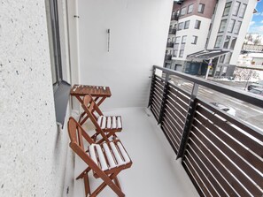 Een balkon met een houten stoel en tafel, een metalen hekwerk en een uitzicht op een stadsbeeld met gebouwen.