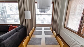 Een moderne woonkamer met een bank, een eettafel met vier stoelen en grote ramen met zonwering.