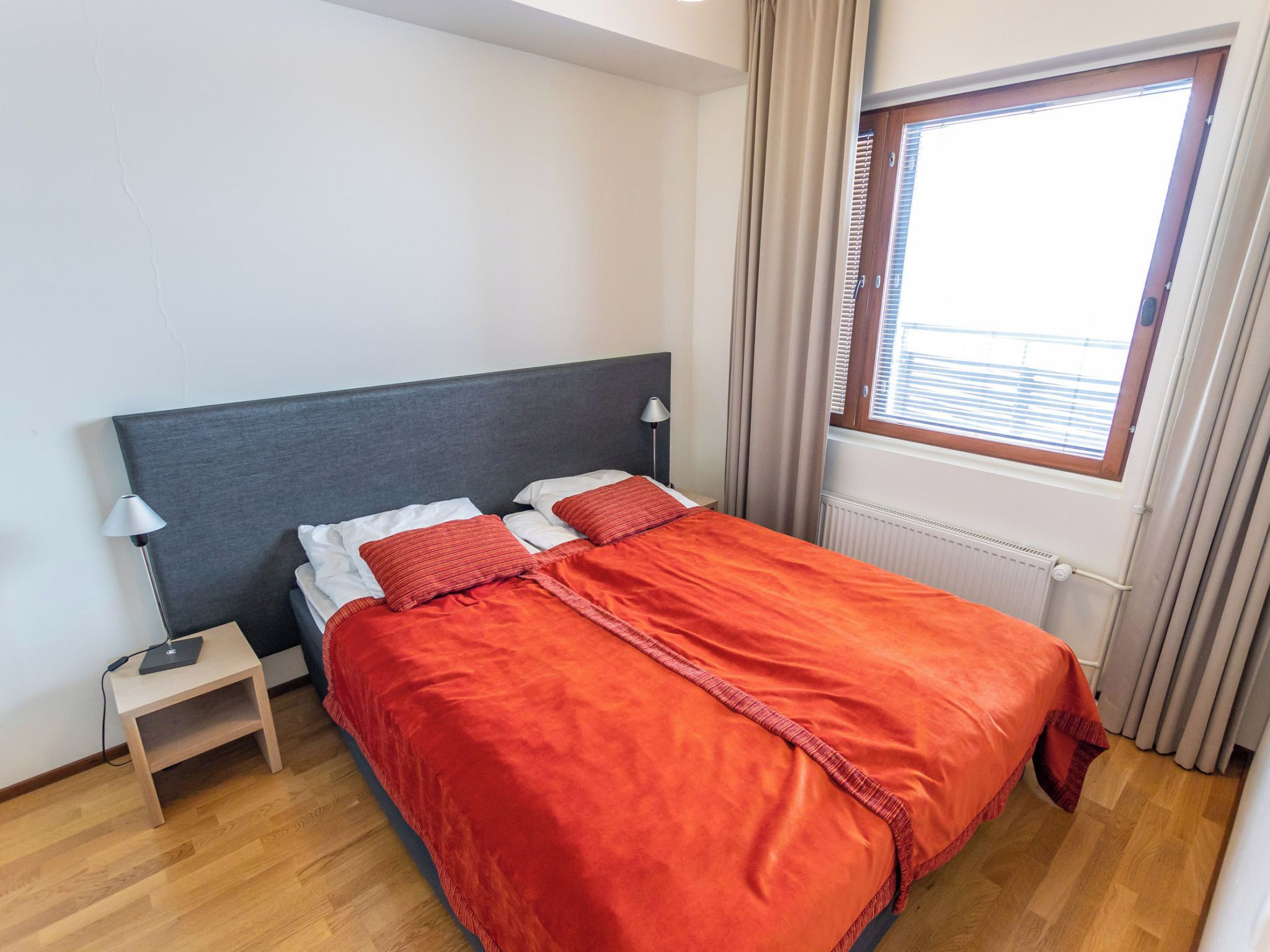 Una habitación de hotel con cama, mesita de noche, lámpara, ventana con persianas y radiador.