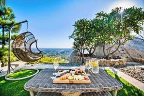 Ein Korbtisch mit einer Platte für Wurst- und Käseplatten, zwei Weingläsern und einem Hängekorbstuhl mit Blick auf eine reizvolle Landschaft.