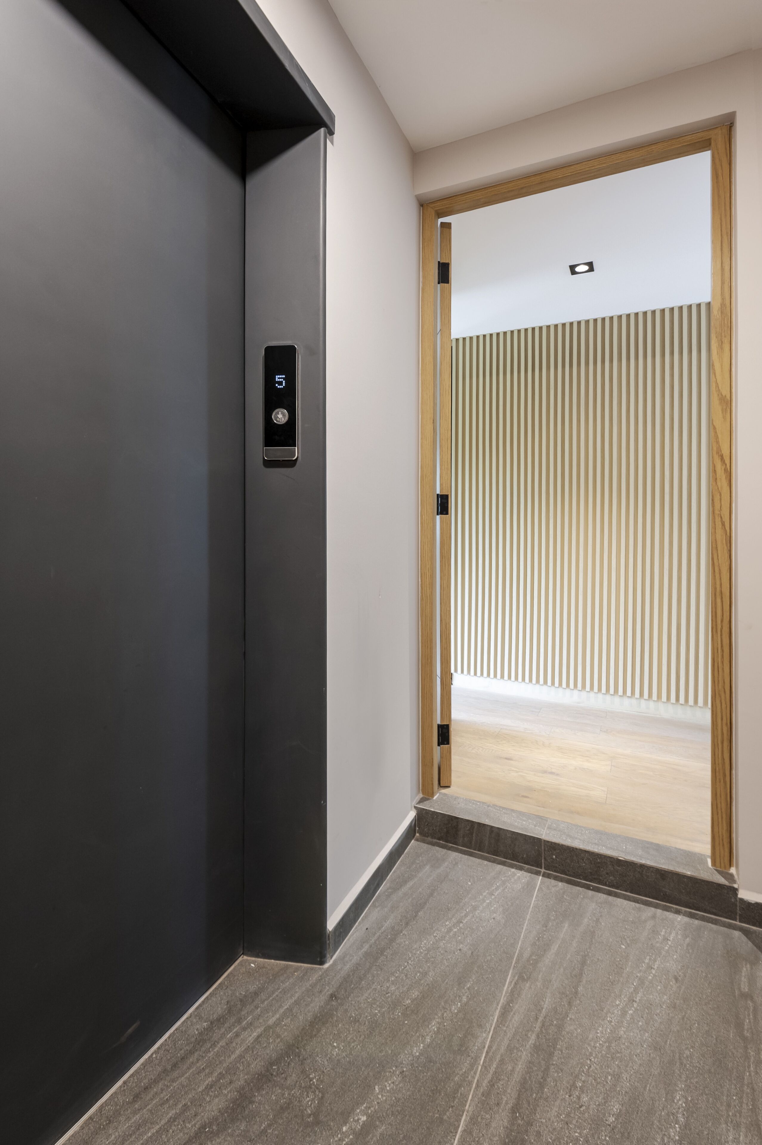 Un ascenseur moderne doté d’un écran numérique et d’une porte à cadre en bois, qui donne accès à une pièce avec un mur à lattes et un plancher en bois.