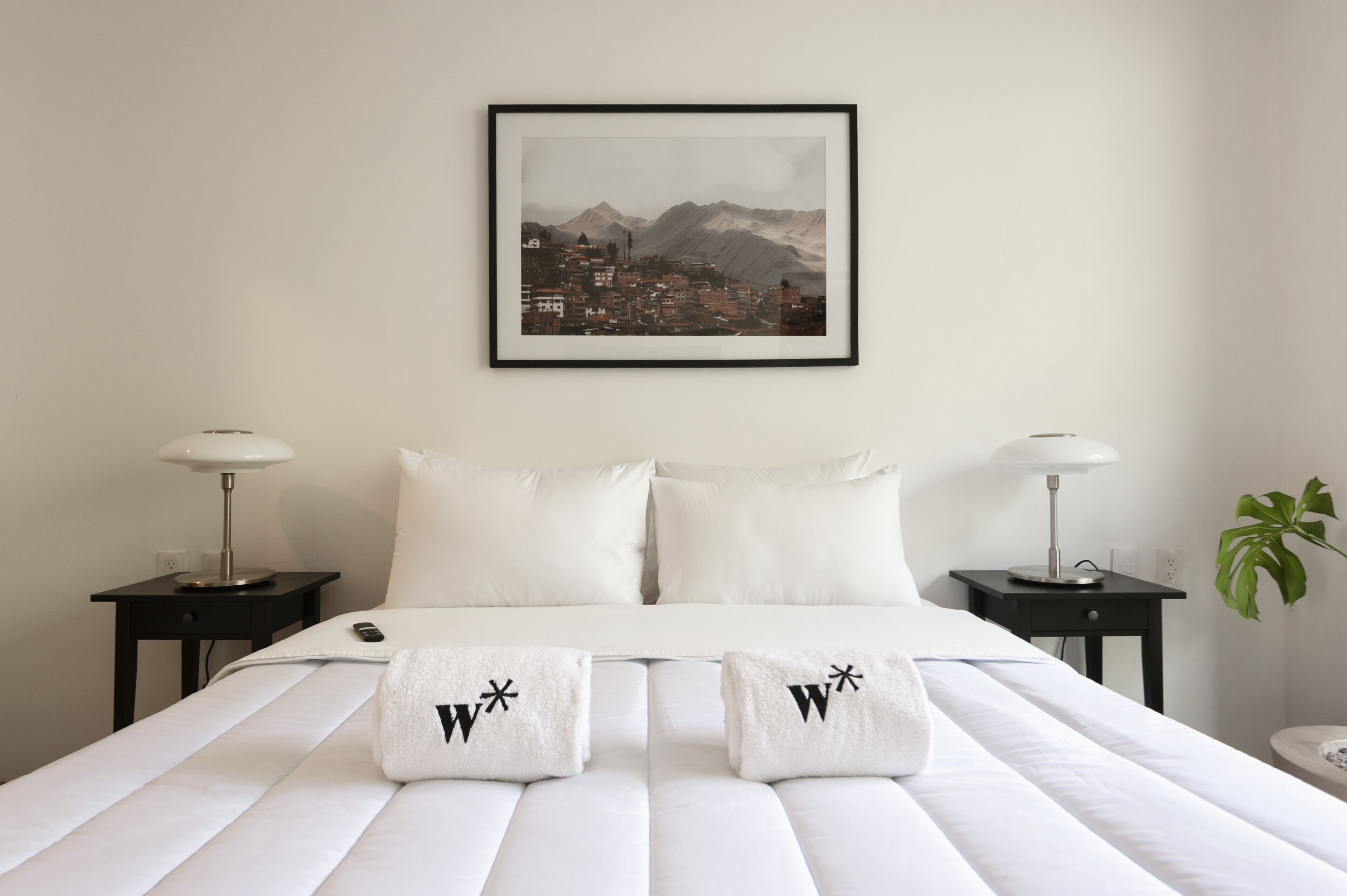 A hotel room with a bed, two nightstands, a framed picture of a mountain town, and a plant.