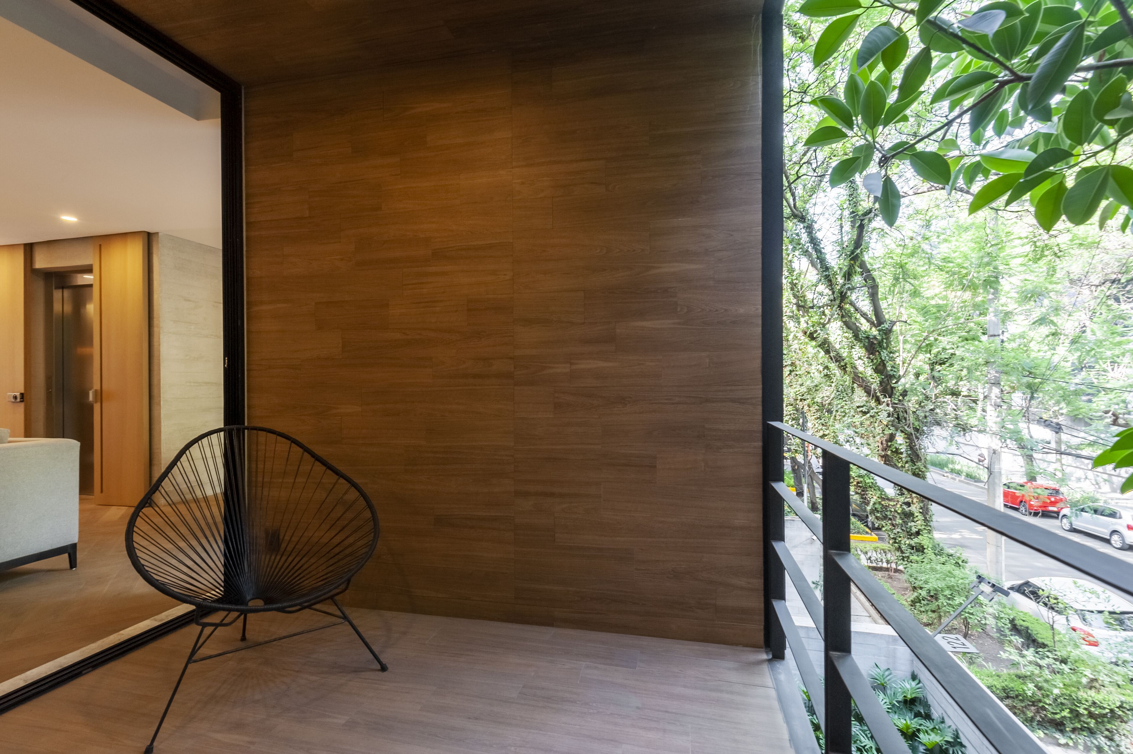 Ein moderner Balkon mit Holzboden, einem schwarzen Drahtstuhl und Blick auf eine Straße mit Autos und Grünflächen.