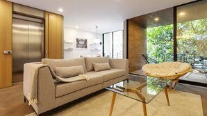 Ein modernes Wohnzimmer mit einer Couch, einem Glastisch, einem Rattan-Sessel, einem großen Fenster mit Blick auf Grünflächen und einem an der Wand hängenden Kunstwerk.