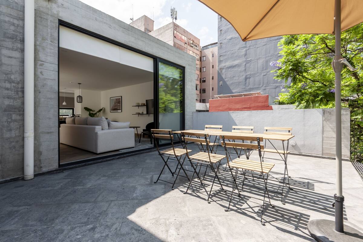 Un patio exterior con un comedor y un sofá, un gran ventanal corredero que da a un espacio interior y vistas de edificios circundantes.
