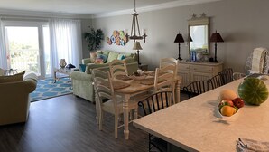 A spacious living room with a dining area, a large window with curtains, a chandelier, and a fruit bowl on the dining table. - Family-friendly beach condo, Indian Beach, NC (Salter Path)