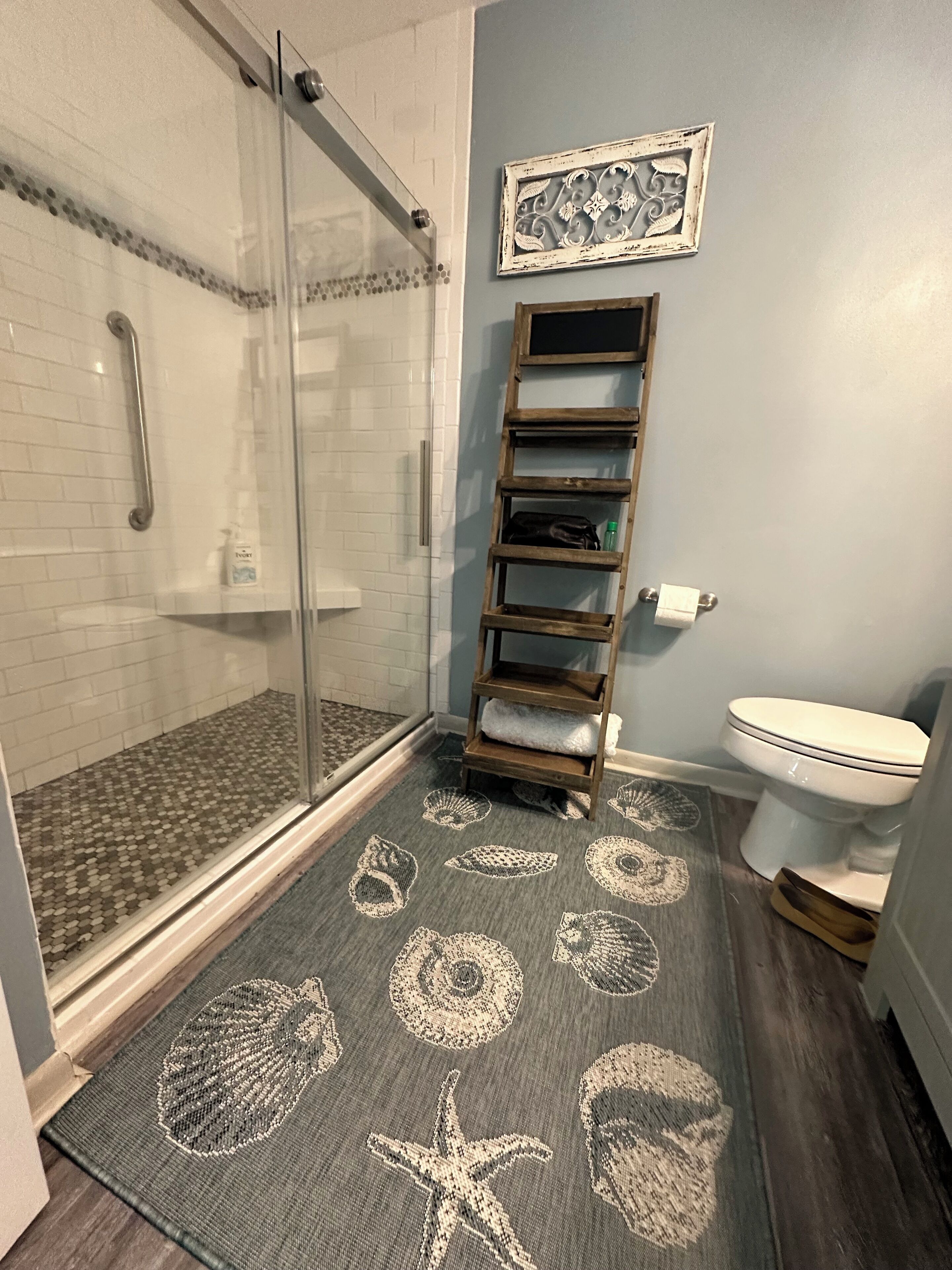 Ein Badezimmer mit Duschkabine aus Glas, einer Holz-Leiterregal, einer Toilette und einem Teppich mit Meeresmotiven.