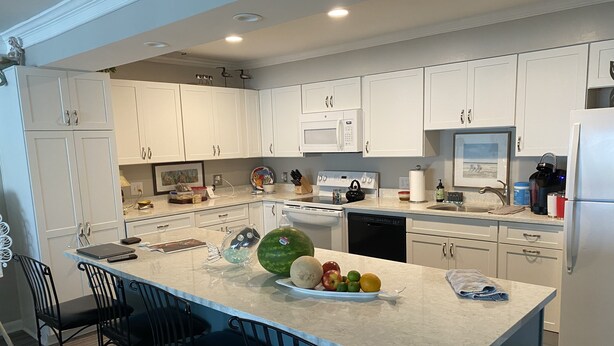 A modern kitchen with white cabinets, a central island with bar stools, and various appliances including a dishwasher and microwave. - Family-friendly beach condo, Indian Beach, NC (Salter Path)