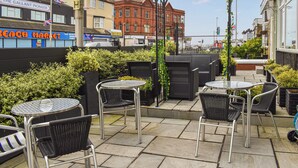 An outdoor patio area with round tables and black wicker chairs, surrounded by greenery and a pergola with climbing plants.