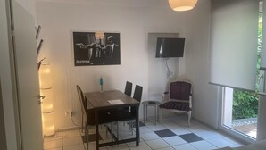 Een kamer met een geruit vloerkleed, een eettafel met stoelen, een aan de muur bevestigde televisie en een raam met uitzicht op groen.