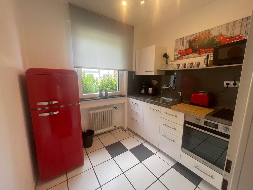Eine kompakte Küche mit einem roten Kühlschrank, weißen Schränken, einem schwarz-weißen Fliesenboden, einem Fenster mit Jalousien und einer Mikrowelle über dem Backofen.