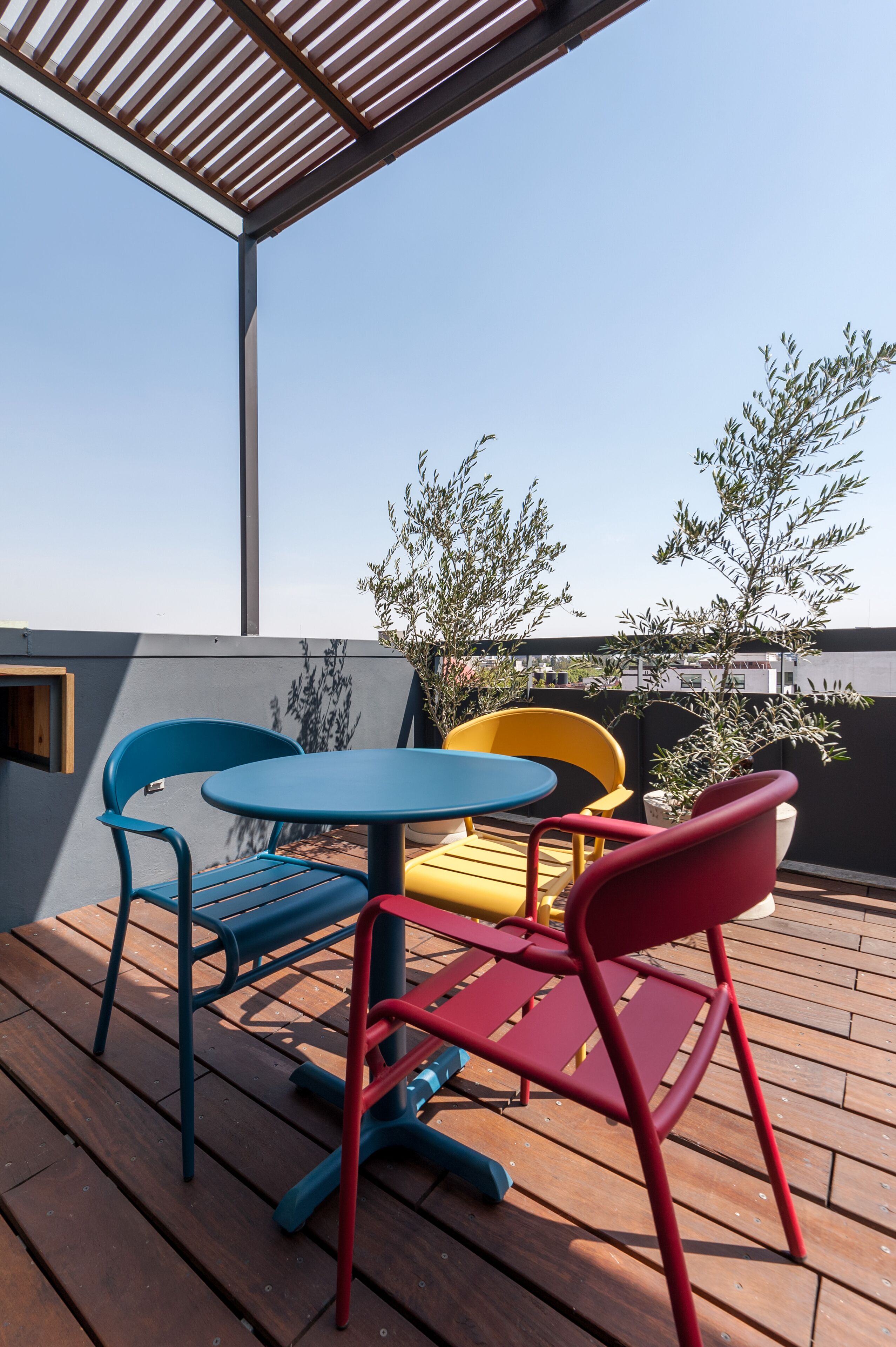 A wooden deck with a table and chairs, a pergola overhead, and olive trees in the background.
