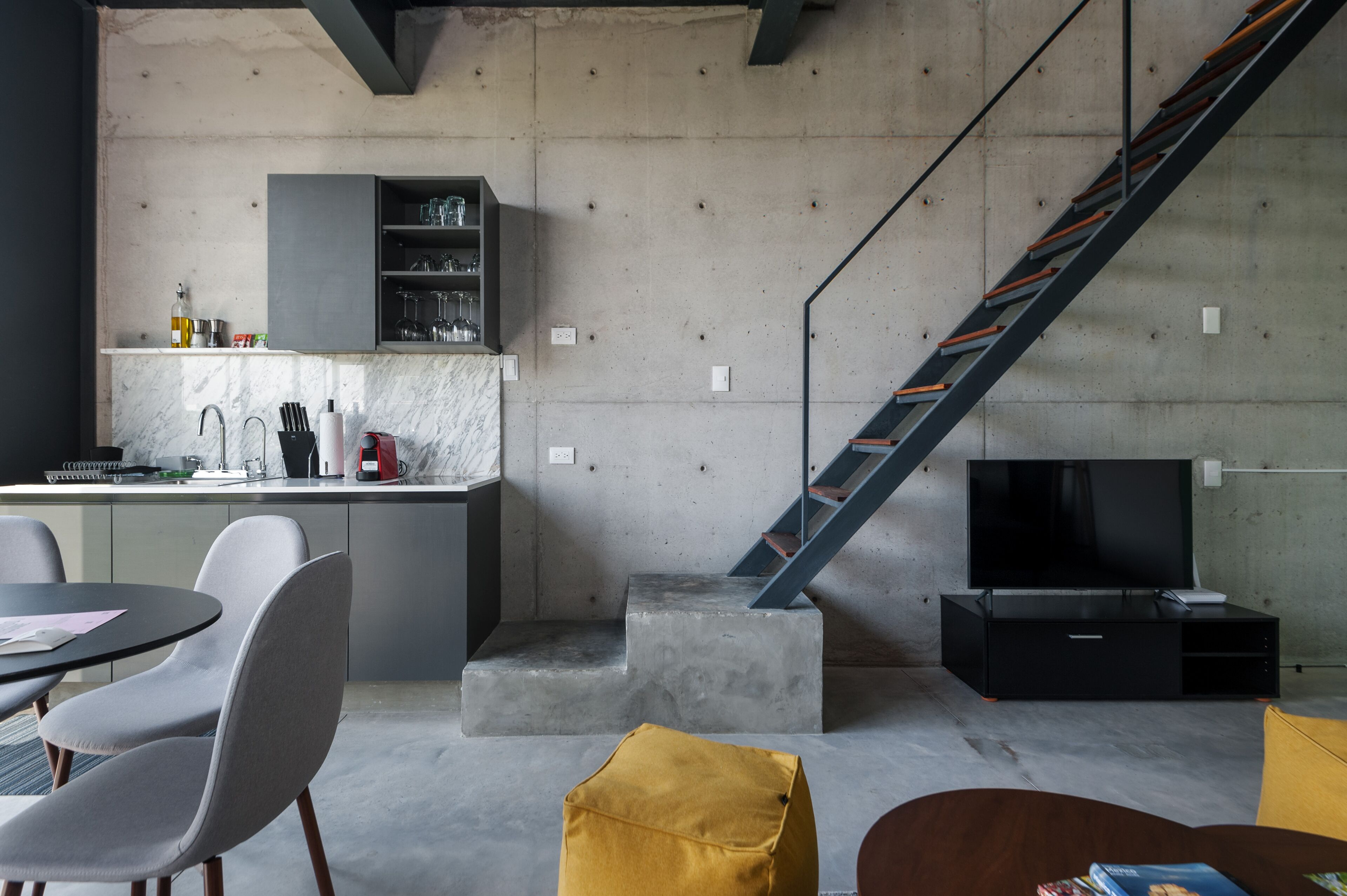 A modern kitchen with a built-in shelf, a staircase, a dining area with chairs, and a flat-screen TV.