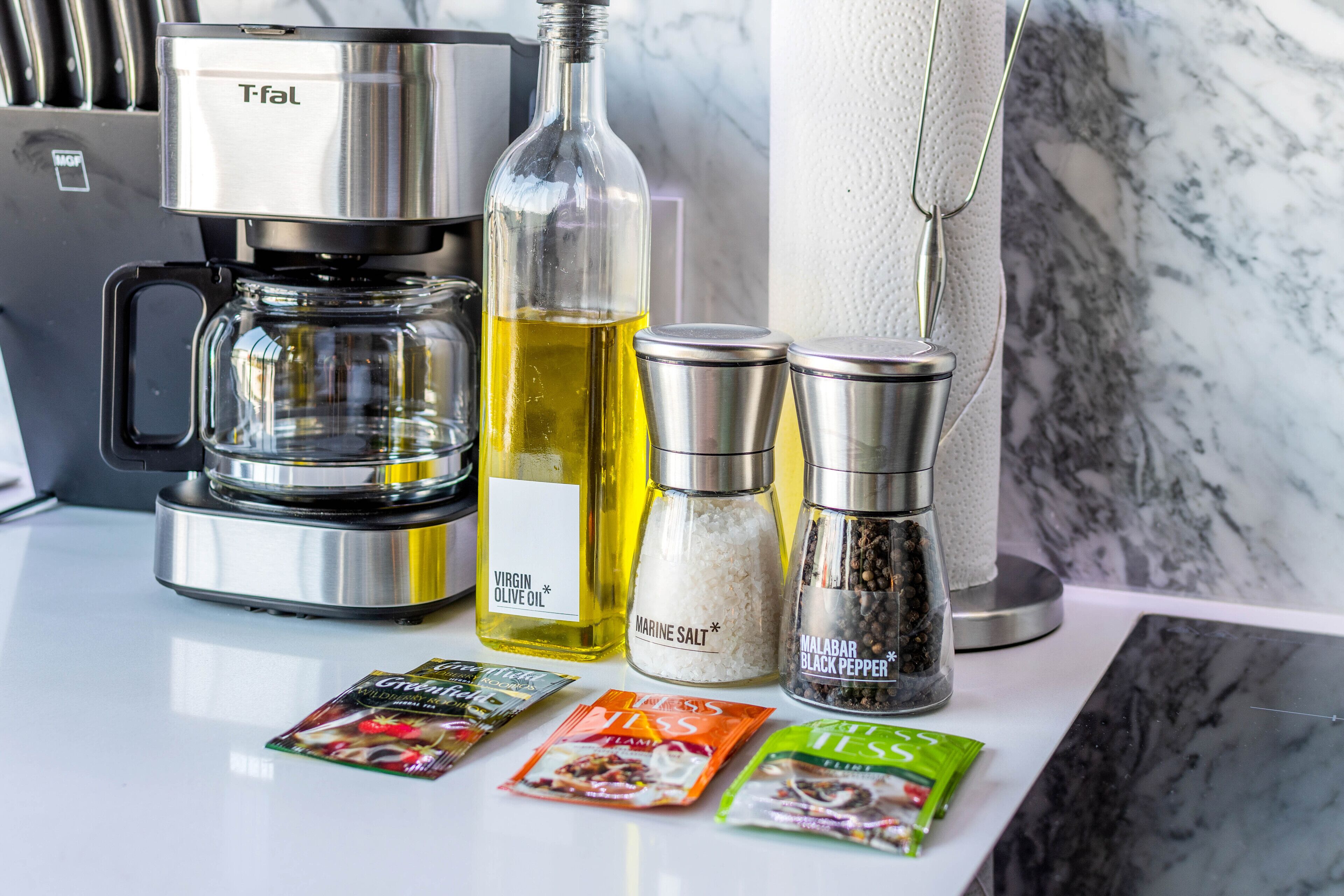 A kitchen counter with a coffee maker, a bottle of olive oil, a container of salt, a container of black pepper, and packets of tea and coffee.