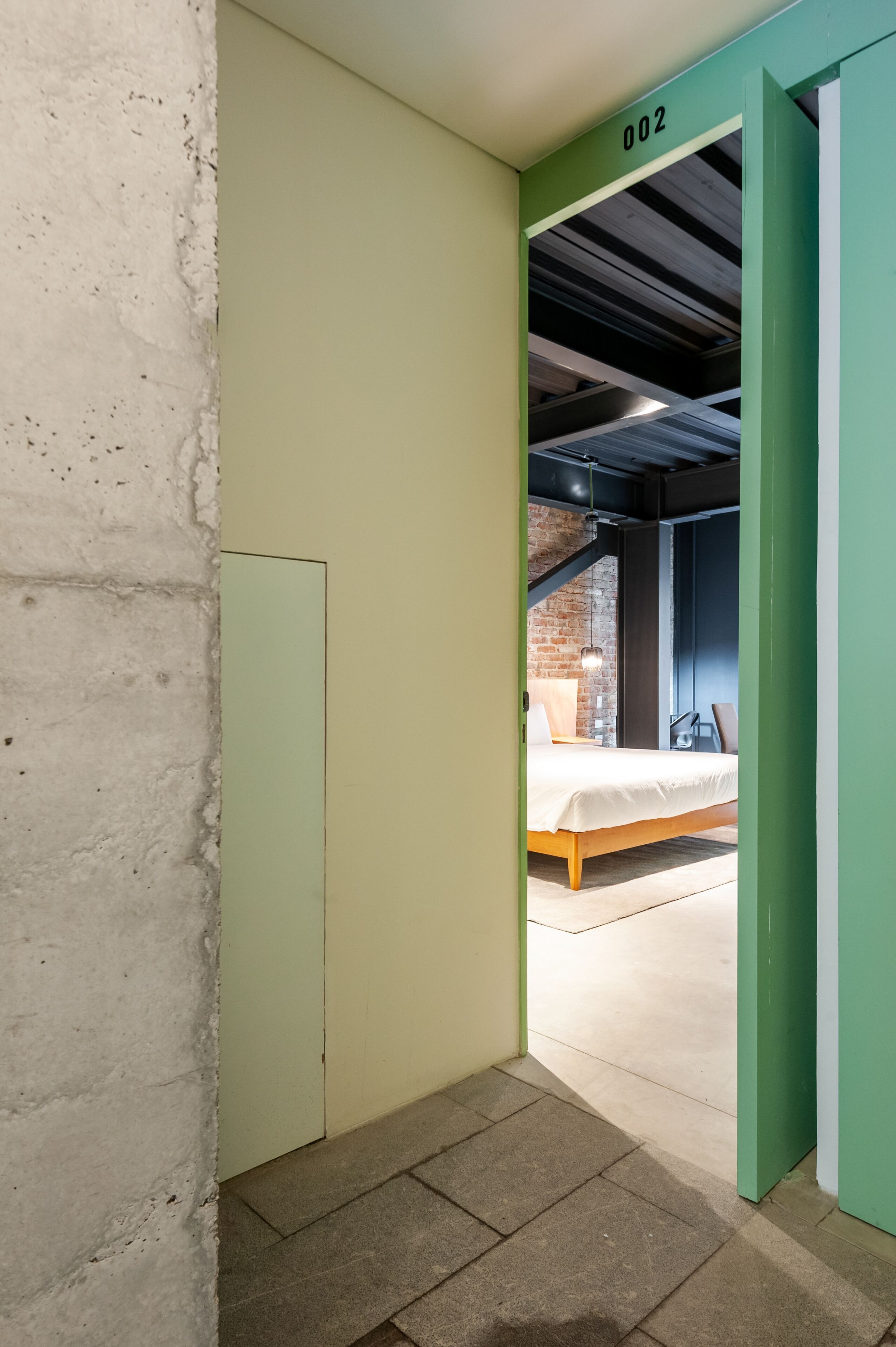 A room with a bed, a brick wall, and a green door with the number 002.