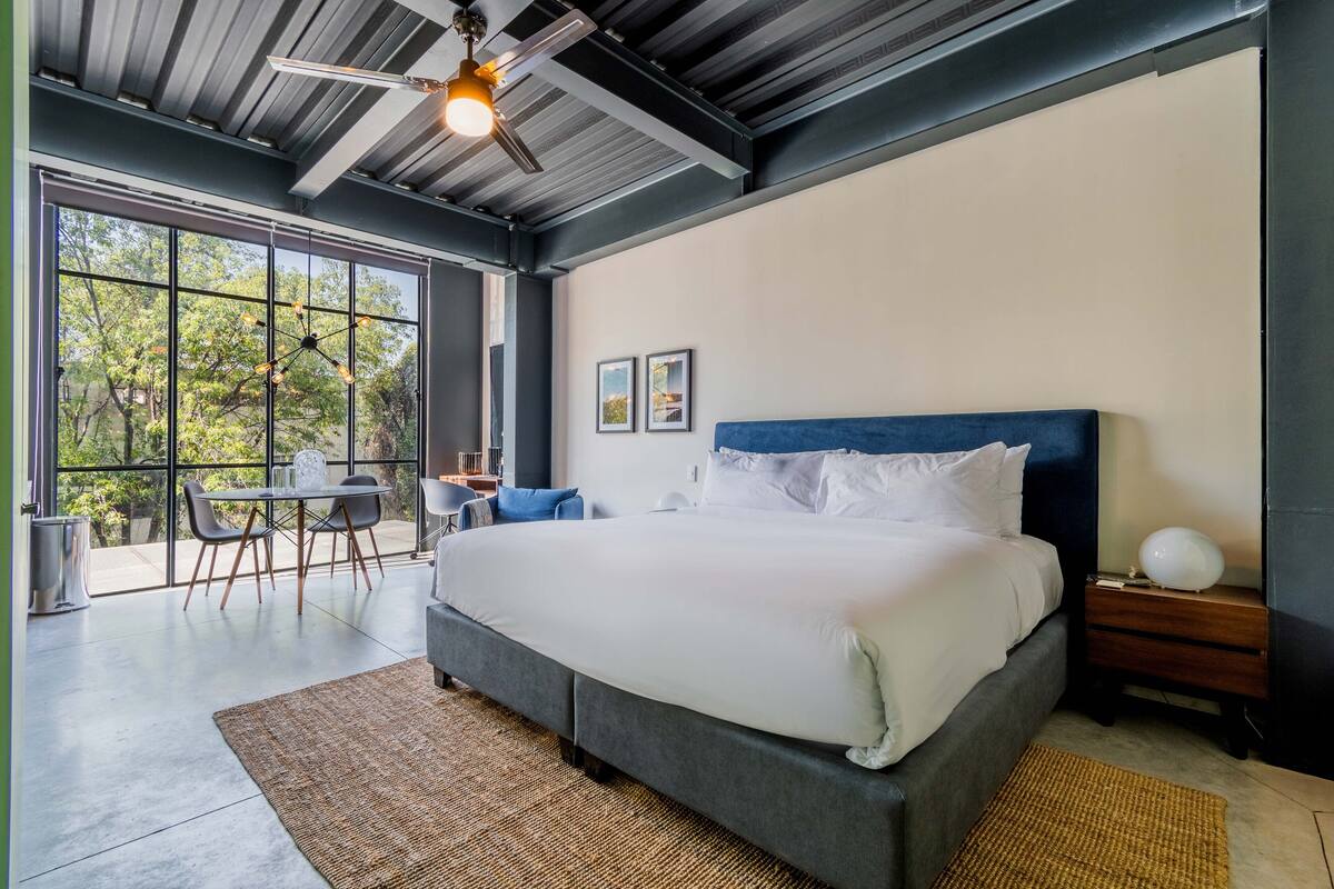 Quarto moderno com uma cama grande, uma mesa de jantar e cadeiras perto de uma janela ampla.
