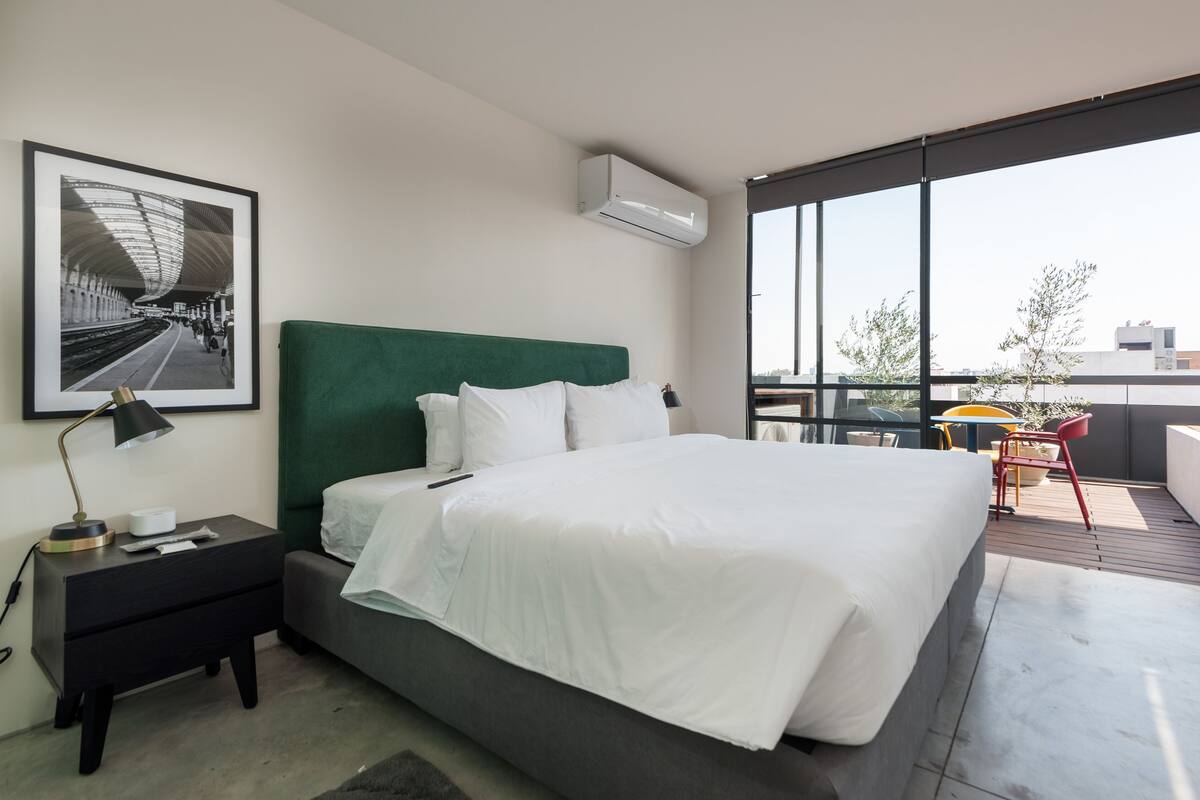 Quarto de hotel moderno com uma cama grande, cabeceira verde, quadro emoldurado na parede, mesinha de cabeceira com abajur, e varanda com mesa e cadeiras.