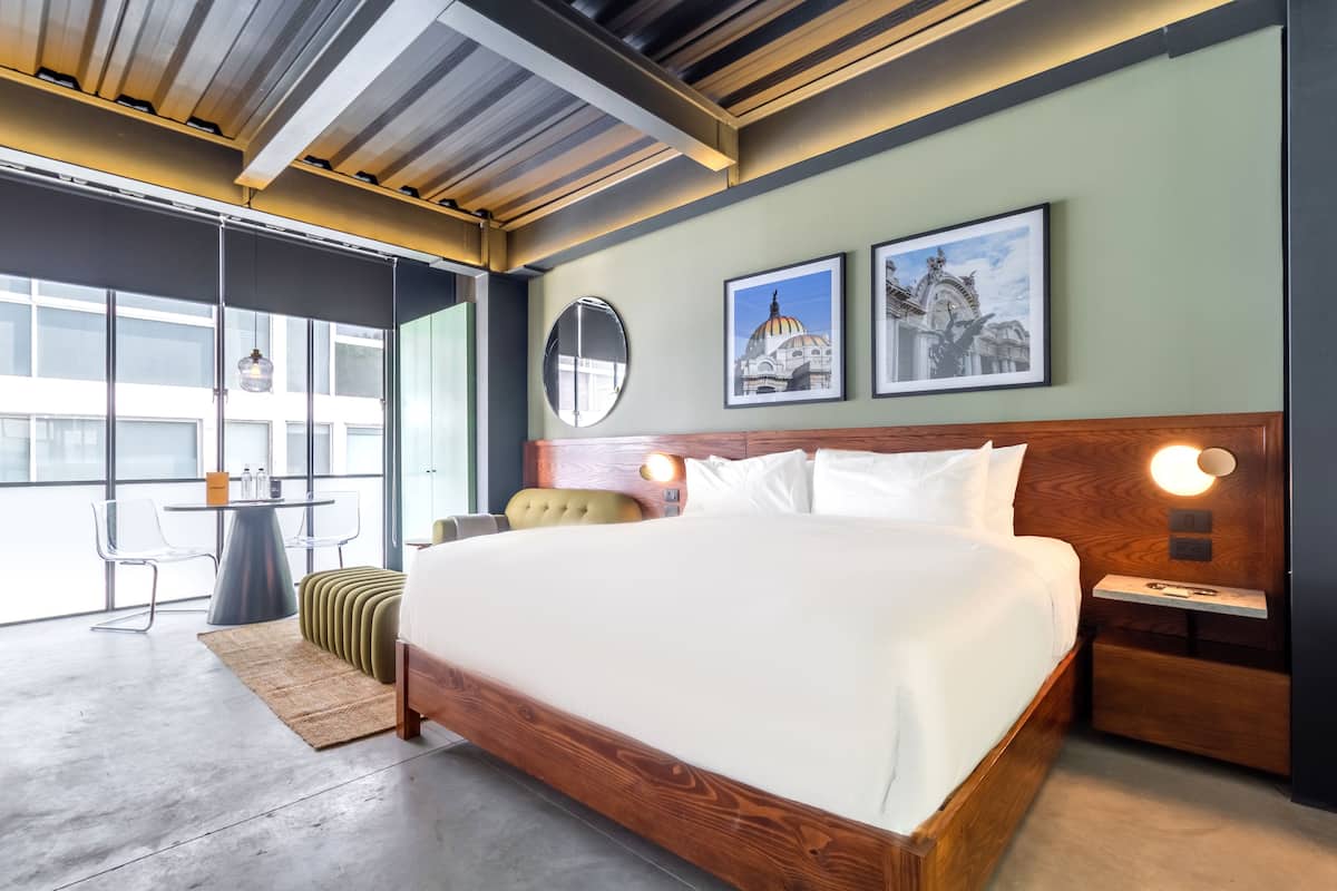 Quarto de hotel moderno com uma cama grande, cabeceira de madeira, mesas de cabeceira, um espelho redondo e obras de arte na parede.
