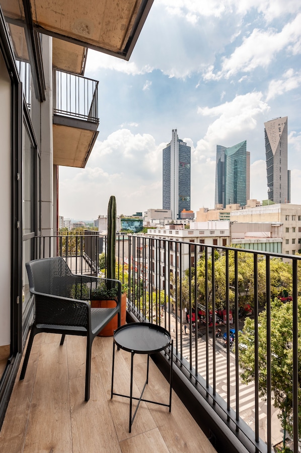 A balcony with a chair, table, and potted plant. It overlooks a cityscape with modern buildings and a cactus.