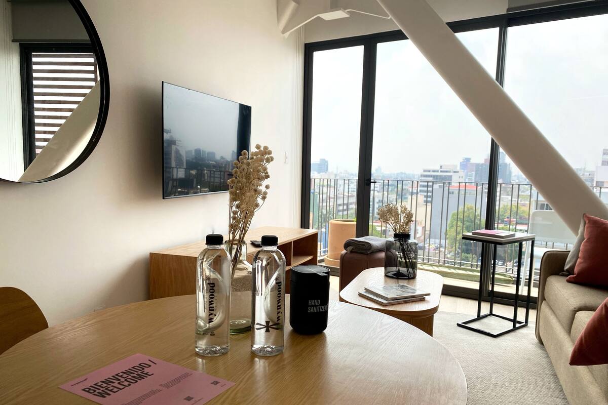 Un salón moderno con una mesa redonda de madera, un sofá y vistas de la ciudad a través de amplias ventanas.