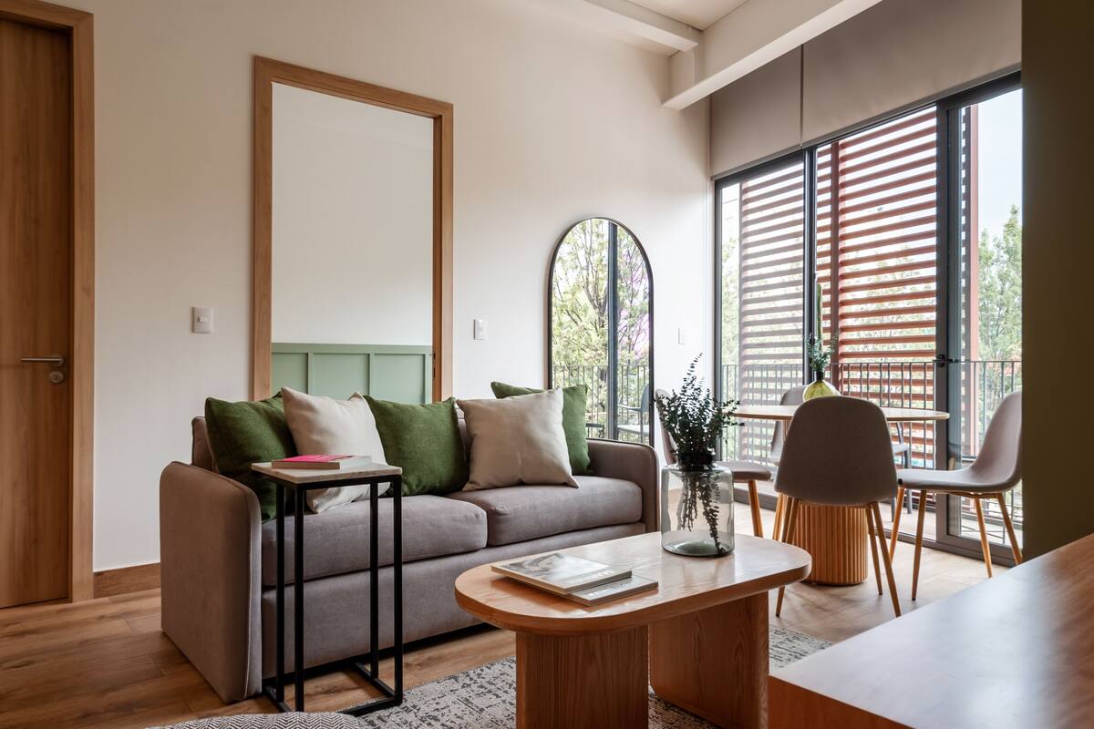 Una sala de estar moderna con un sofá, una mesa de centro y sillas. Hay un ventanal con persianas, un espejo y una puerta.