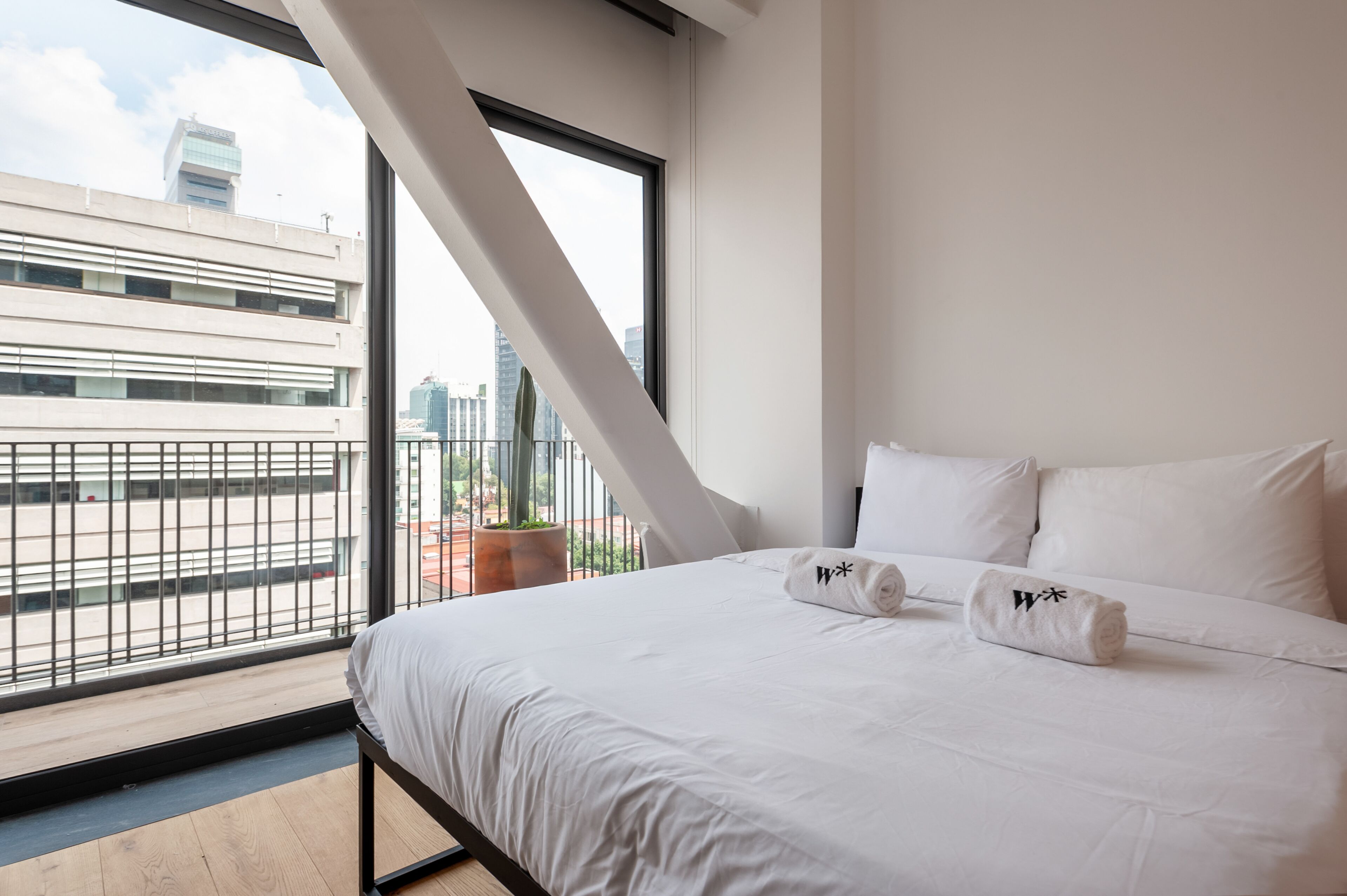 A modern hotel room with a large bed, white linens, and a view of the city through large windows.