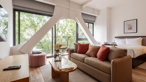 A modern hotel room with a large bed, a sofa, a coffee table, and a desk. There are large windows offering a view of trees and a balcony. - Wynwood House Londres (Mexico City)
