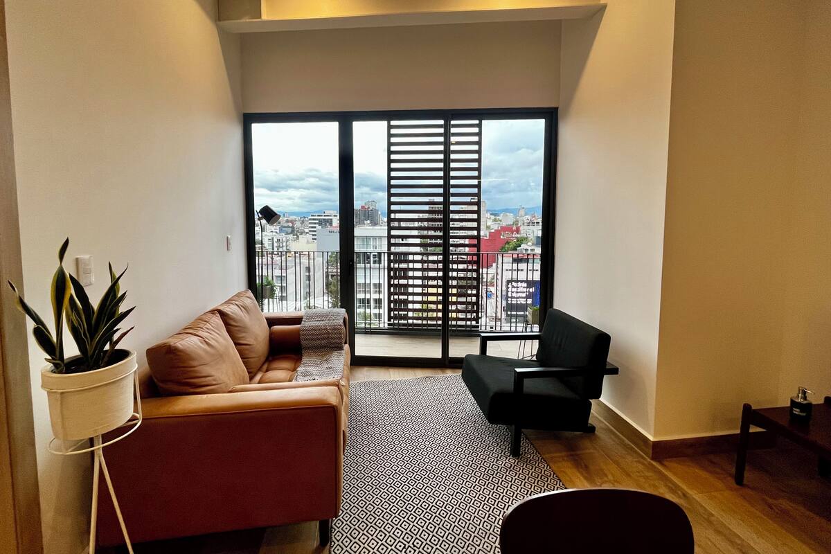 Una sala de estar moderna con un sofá, un sillón negro, un tapete estampado y una planta en maceta.